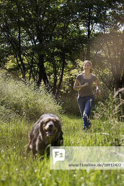 Junge Frau rennt ihrem Hund auf einer Wiese hinterher.