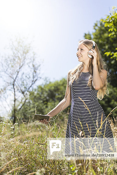 Lächelnde junge Frau steht auf der Wiese und hört Musik mit Smartphone und Kopfhörer.