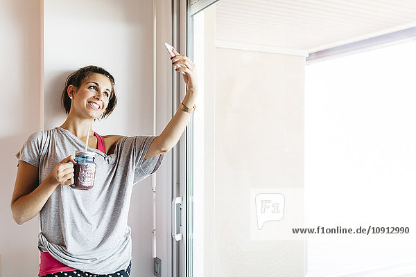 Lächelnde junge Frau mit gesundem Getränk  die Selfie am Fenster nimmt.