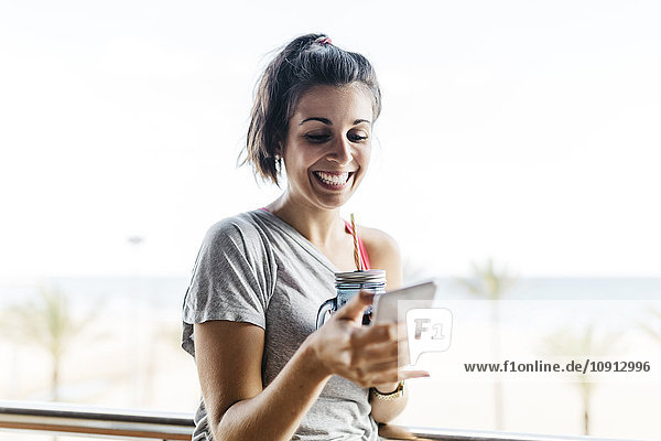 Happy young woman with healthy drink and smartphone on balcony