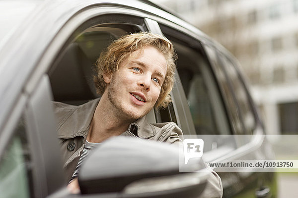 Porträt eines lächelnden jungen Mannes  der sich aus dem Autofenster lehnt und etwas beobachtet.