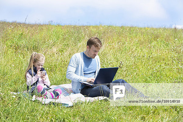 Vater und Tochter sitzen auf der Wiese  Smartphone und Laptop