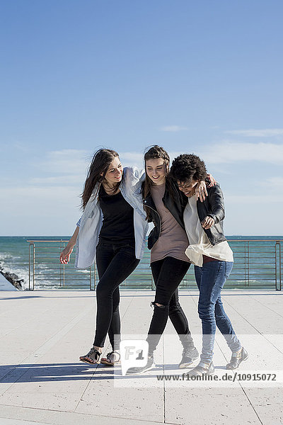 Drei junge Frauen gehen Arm in Arm am Meer.