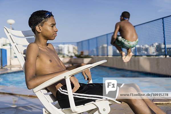 Teenager-Junge sitzt auf einem Stuhl vor dem Schwimmbad  während sein Freund einen Kanonenkugel-Tauchgang macht.