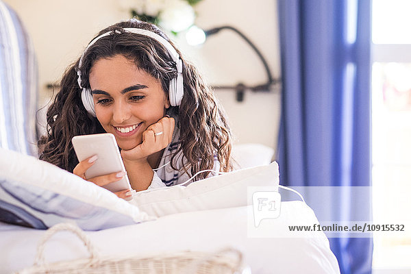 Lächelndes Teenagermädchen auf der Couch liegend mit Smartphone und Kopfhörer