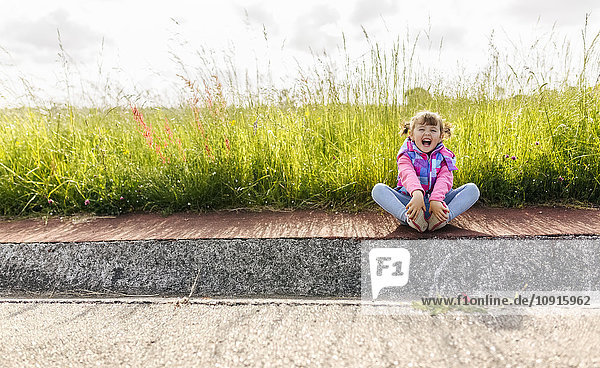 Lachendes kleines Mädchen auf dem Bürgersteig vor einer Wiese sitzend