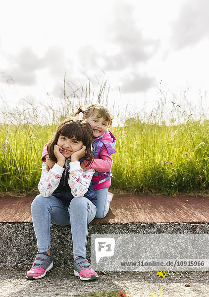 Porträt von zwei kleinen Schwestern  die auf dem Bürgersteig vor einer Wiese sitzen.