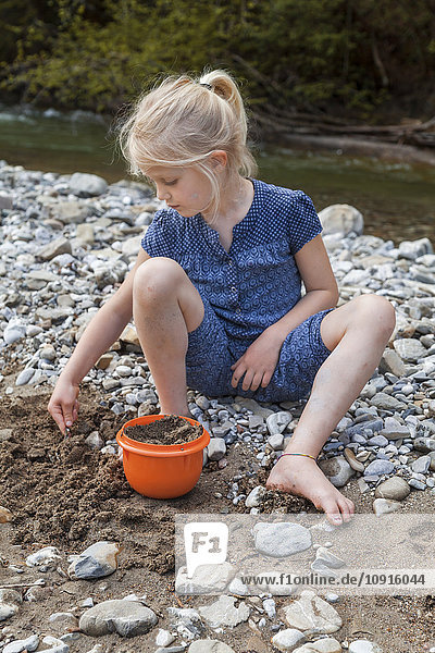 Blondes kleines Mädchen spielt mit Sand am Flussufer