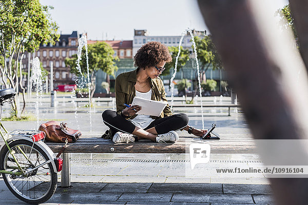 Junge Frau sitzt auf einer Bank mit Notizbuch und schaut auf ein digitales Tablett.