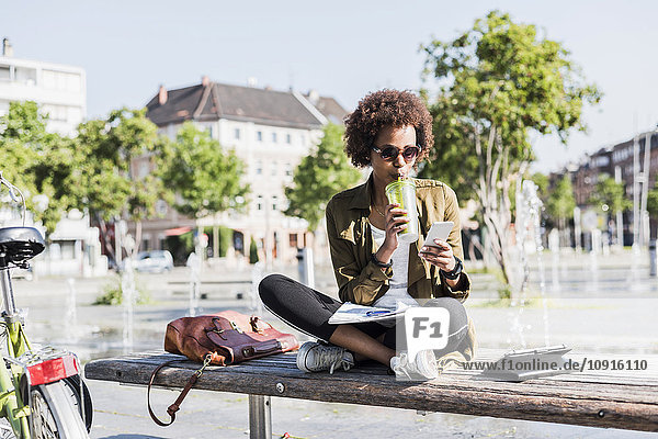 Junge Frau sitzt auf einer Bank und trinkt ein Getränk  während sie auf ihr Smartphone schaut.