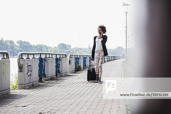 Lächelnde Geschäftsfrau mit Gepäcktelefonieren mit Smartphone am Flussufer
