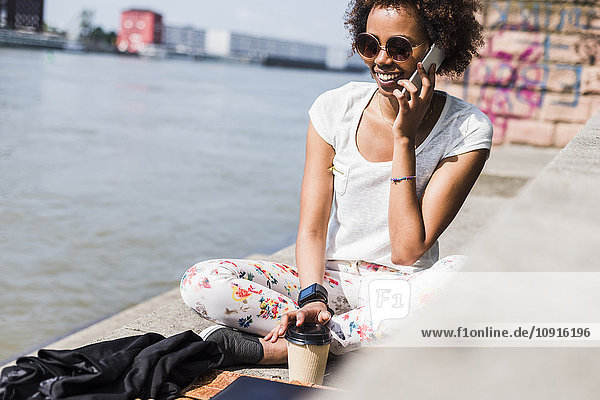 Junge Frau mit Kaffee zum Telefonieren mit Smartphone am Flussufer