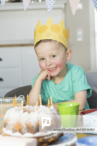 Glücklicher Junge mit Papierkrone am Geburtstagstisch sitzend