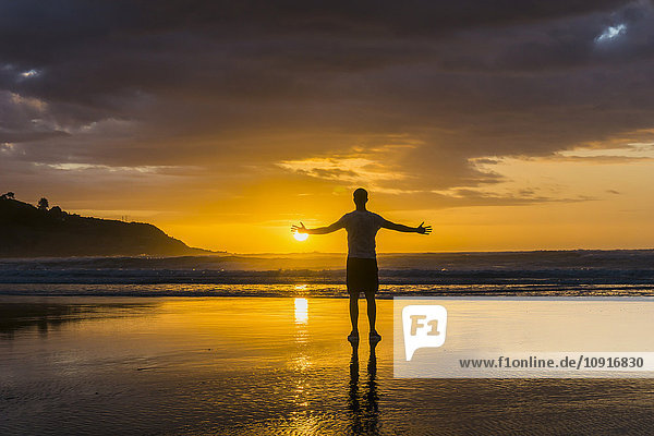 Silhouette des am Strand stehenden Mannes mit ausgestreckten Armen und Blick aufs Meer