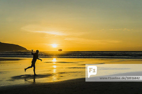 Silhouette eines Mannes am Strand  der in der Dämmerung mit Frisbee spielt.