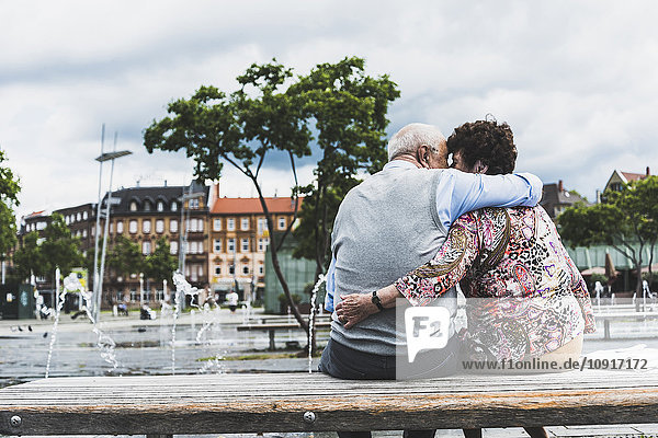 Deutschland  Mannheim  Rückansicht eines küssenden Seniorenpaares  das auf einer Bank sitzt und sich umarmt.