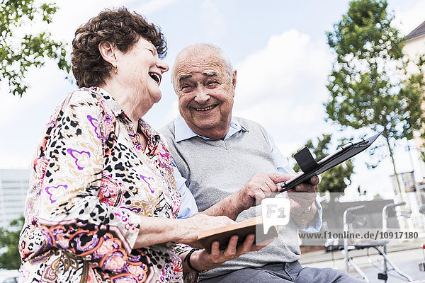Lachendes Seniorenpaar sitzend auf einer Bank mit Buch und Tablette