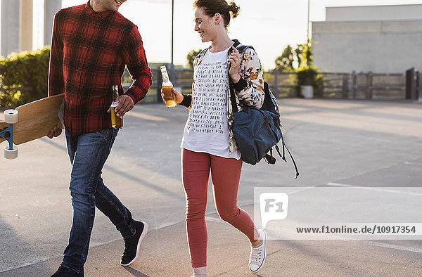 Young couple with beer bottles and skateboard walking outdoors