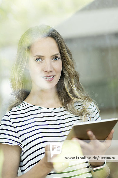 Porträt der lächelnden Frau mit digitalem Tablett hinter der Fensterscheibe