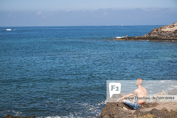 Mann sitzt auf einem Felsen und schaut zum Meer.