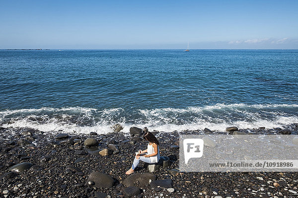 Frau mit Notizbuch am steinigen Strand am Meer sitzend