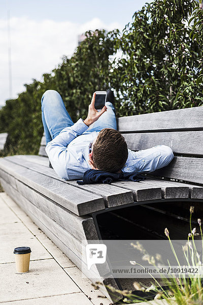 Junger Mann auf der Bank liegend mit Smartphone