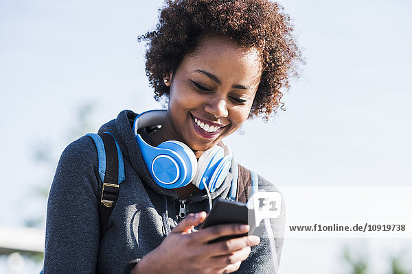 Smiling young woman wearing headphones looking at cell phone outdoors