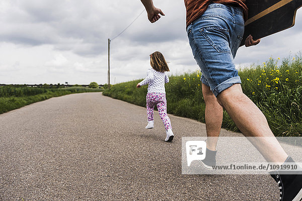 Father with skateboard and daughter running on country lane