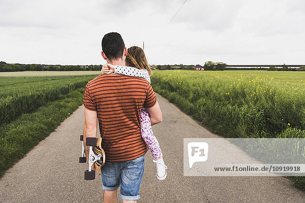 Father with skateboard carrying daughter on country lane