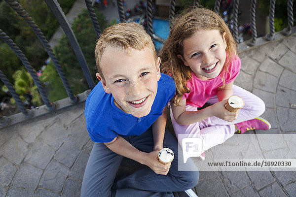 Portrait of brother and sister with ice cream cones sitting on balcony looking up to camera