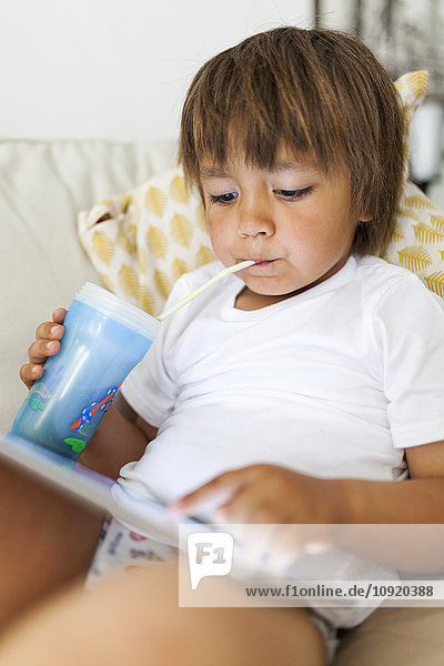 Litte boy sitting on couch using digital tablet while drinking something