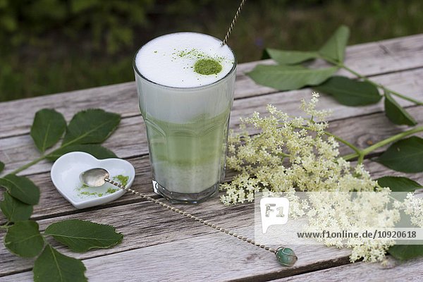 Matcha-Tee mit Milch im Glas auf Holz  Holunderblüten und herzförmiger Porzellanschale mit Löffel