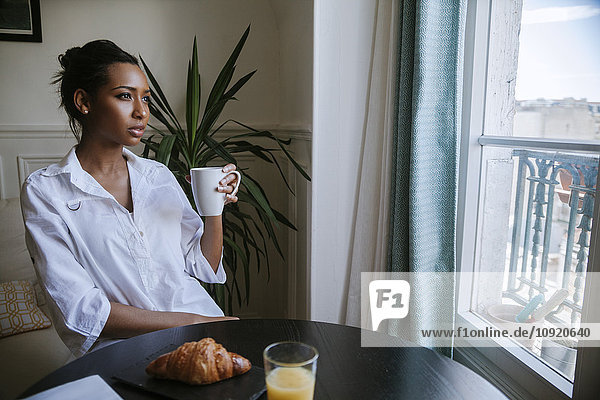Junge Frau beim Frühstück mit einer Tasse Kaffee und Blick durchs Fenster