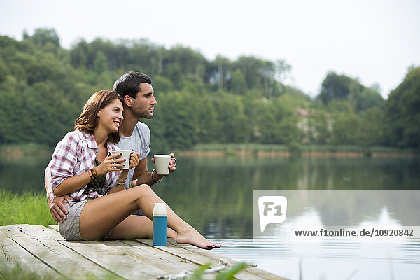 Relaxed young couple with coffee mugs sitting on a jetty at lake looking at distance
