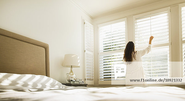 Frau im Bademantel öffnet Fensterläden im Schlafzimmer