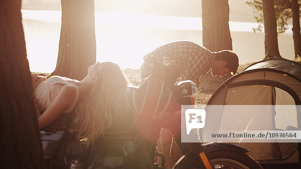 Junger Mann bereitet Zelt auf Campingplatz neben Frau auf Motorrad liegend vor