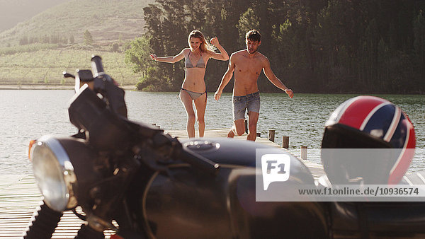 Junges Paar  das auf einem Steg am Seeufer in Richtung Motorrad läuft