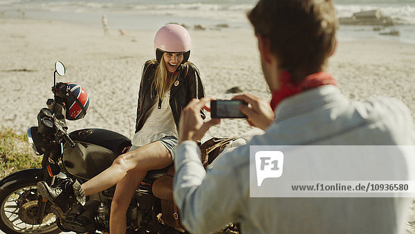 Junger Mann fotografiert Frau auf Motorrad am Strand