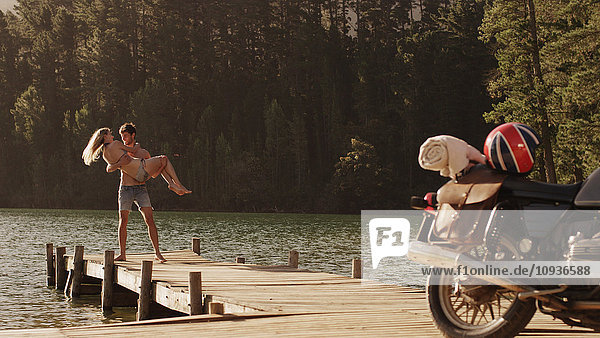Junger Mann trägt junge Frau auf einem Steg am See in der Nähe eines Motorrads