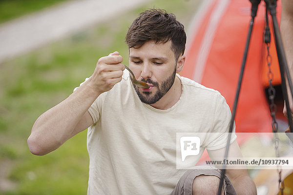 Young man at campsite tasting food with spoon