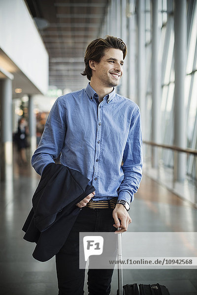 Happy businessman with luggage looking away while standing at airport