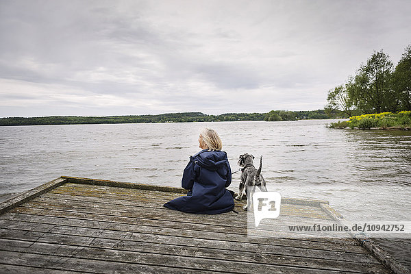 Rear view of senior woman and dog on pier at lake against cloudy sky