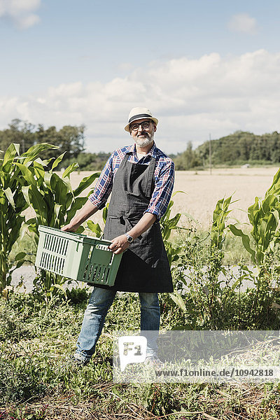 Portrait of smiling mature gardener holding crate and standing at farm