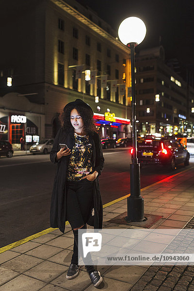 Volle Länge der Frau,  die das Smartphone benutzt,  während sie nachts auf dem Bürgersteig in der Stadt steht.