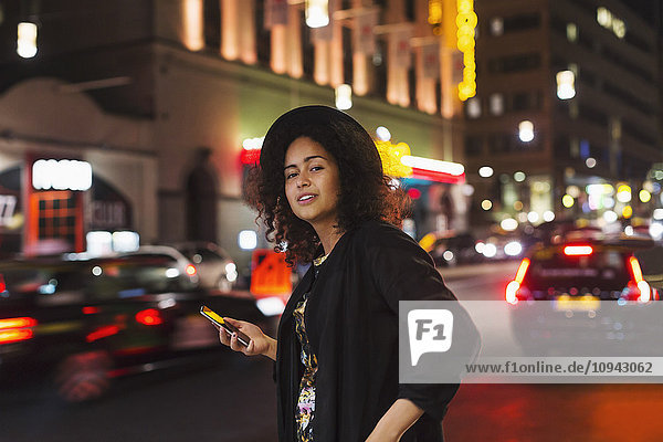 Porträt einer selbstbewussten Frau mit Smartphone in der Stadt bei Nacht