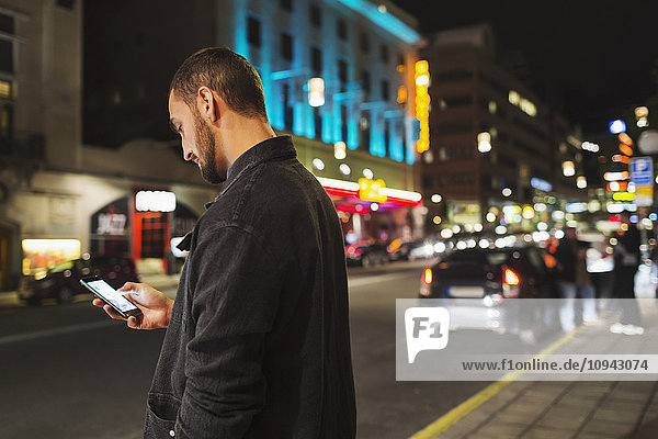 Seitenansicht des Mannes mit dem Smartphone auf der Stadtstraße bei Nacht