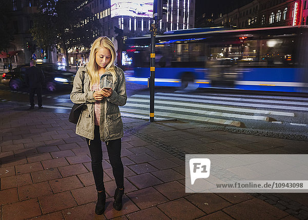 Volle Länge der Frau mit dem Handy auf dem Bürgersteig in der Stadt in der Nacht