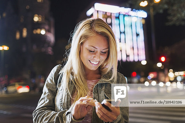 Fröhliche junge Frau mit Smartphone auf der Straße in der Stadt bei Nacht