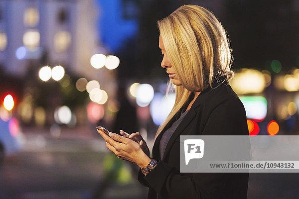 Seitenansicht der jungen Frau mit Smartphone auf der Stadtstraße bei Nacht