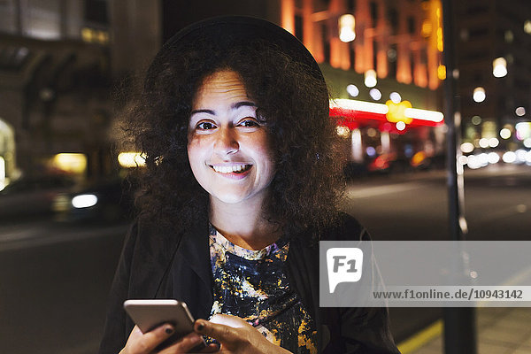 Porträt einer lächelnden Frau  die beißt  während sie nachts ein Smartphone in der Stadt hält.